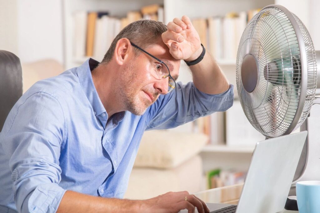 man who is very hot and sweaty sits at a desk in front of a computer with a fan blowing on him